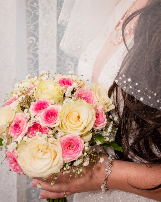 A bride clutching a floral bouquet consisting of large, creamy white roses and smaller, vibrant pink roses, complemented by sprigs of delicate white baby's breath flowers. The bouquet is wrapped in wh
