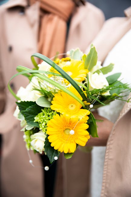 A floral arrangement featuring bright yellow gerbera daisies, white roses, and green foliage, including long, slender leaves and textured filler greenery. The bouquet is held by a person wearing a bei