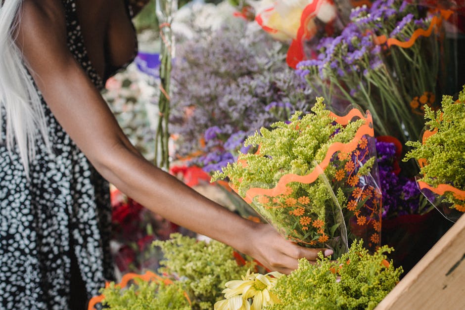 A person's hand holding a vibrant bouquet of fresh yellow and green flowers wrapped in clear plastic with orange decorative accents, set against a background of various colorful floral arrangements in