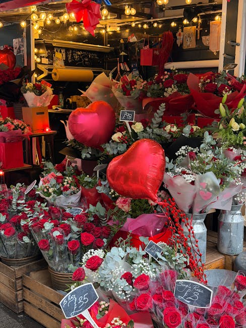 A vibrant display of floral arrangements featuring red and white roses, pink tulips, and baby's breath, arranged in baskets and wrapped in tissue paper. The flowers are set on a rustic wooden table wi