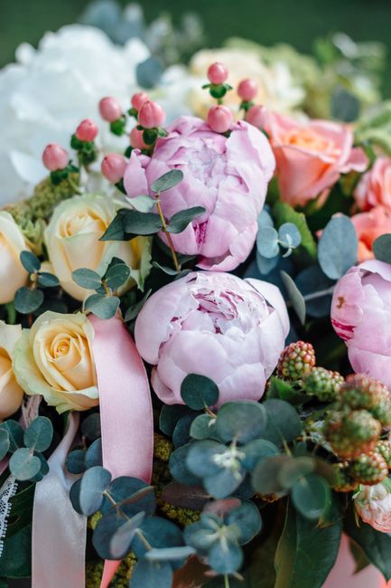 A close-up view of a floral arrangement featuring soft pink peonies with ruffled petals, creamy yellow roses with tightly curled petals, and pink berries with green stems. The bouquet is accented with