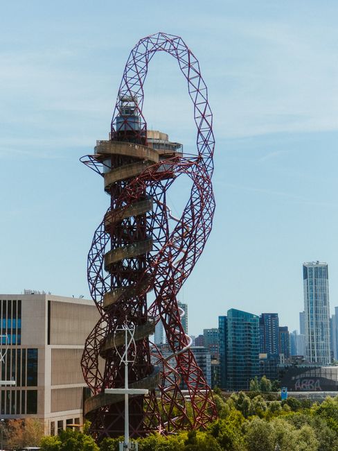 A tall, open steel sculpture resembling a human figure with an ornate, looping head and body, painted in shades of red and purple, situated outdoors in an urban setting with modern high-rise buildings