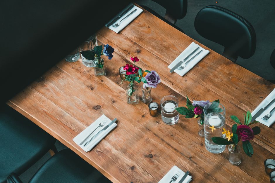 A wooden dining table set for a floral arrangement display features five small glass vases containing fresh flowers in shades of purple, pink, yellow, and blue, arranged along its length. Each vase ho
