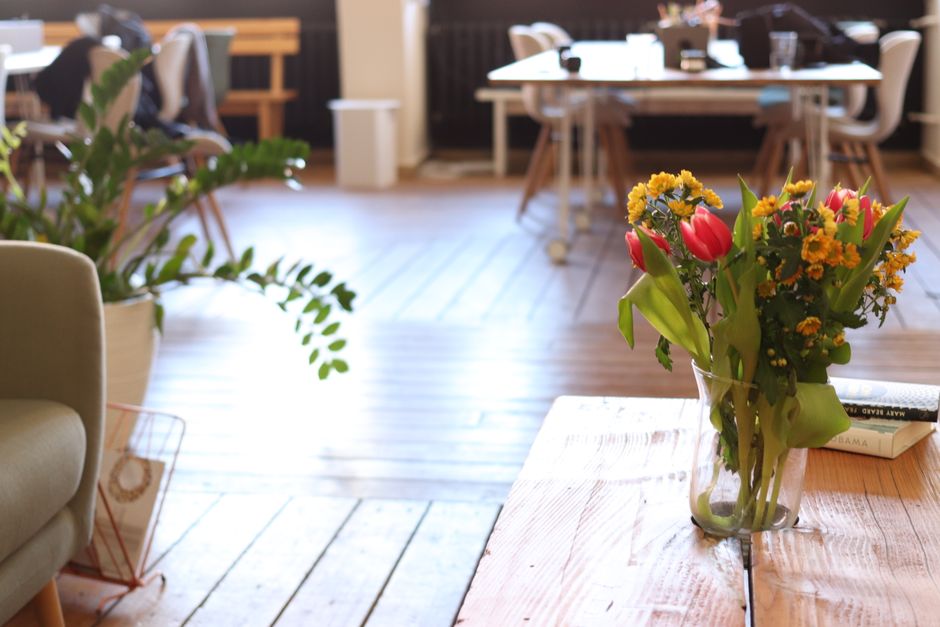 A clear glass vase filled with a fresh floral arrangement consisting of red tulips, yellow daisies, and small orange marigolds, sits on a light wooden table in a well-lit, modern indoor space. The bou