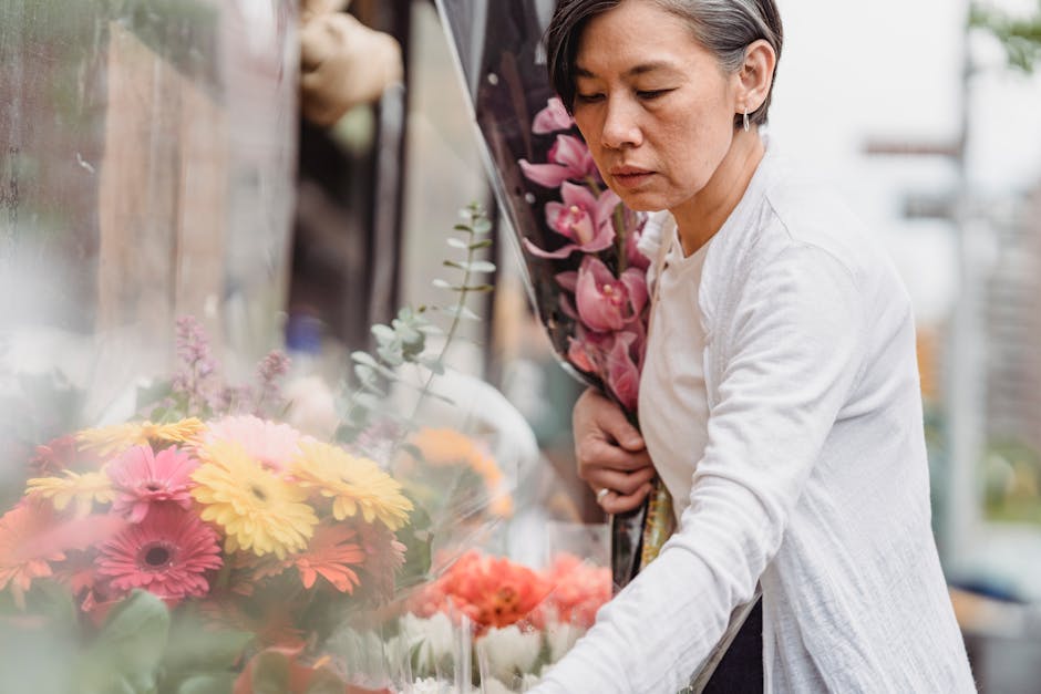 A woman holding a bouquet of pink orchids and standing next to a floral display featuring a variety of fresh flowers, including yellow gerbera daisies, pink gerbera daisies, and orange blooms, arrange