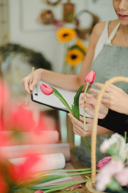 A florist from Flowers Hackney Wick holds a delicate bouquet of fresh pink tulips with smooth, vibrant petals and upright green stems, arranging or inspecting the floral gift at a workspace. The bouqu