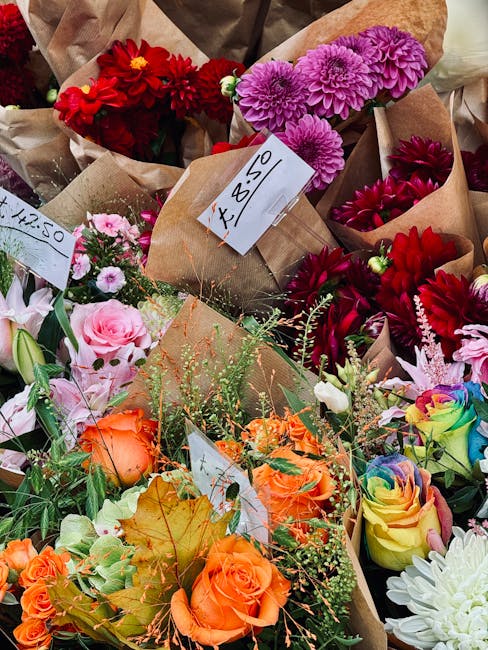 A vibrant collection of flower bouquets arranged on a table, each wrapped in brown kraft paper and labeled with handwritten price tags. The arrangements include deep red dahlias, bright pink chrysanth