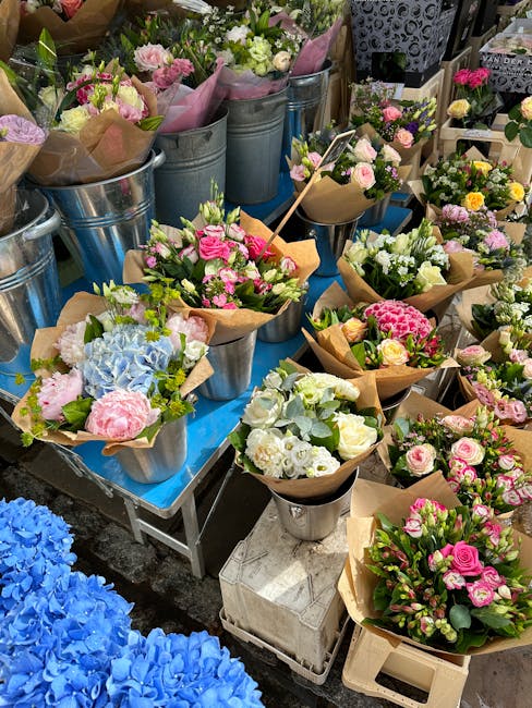A vibrant display of fresh flower bouquets arranged in metal buckets and paper-wrapped bundles, featuring a variety of roses, hydrangeas, carnations, and lisianthus in shades of pink, white, cream, an