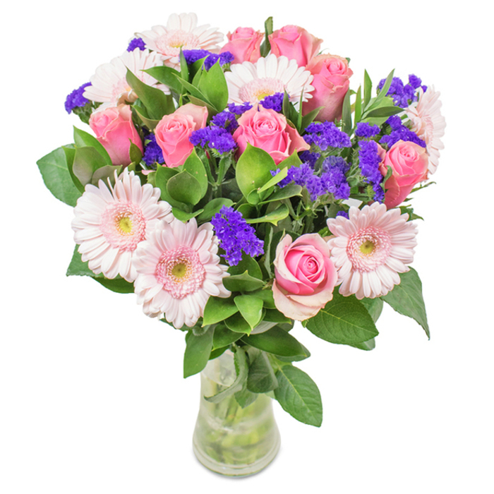 Presented in a clear glass vase and set against a pristine white, this bouquet is a beacon of Hackney Wick charm. The centerpiece showcases seven to nine blushing pink roses, their classic spiral petals opening in slow, graceful unfurling-symbols of joyful affection and gentle romance. Intertwined with these are five to seven large, pale pink gerbera daisies, their bright, nearly white centers providing a lively contrast and a nod to Hackney's playful artistry. Bright purple statice are interspersed throughout, their crisp texture adding energy and modern flair. The bouquet is anchored by a generous spread of deep green foliage, reflecting the abundant trees and parks that define Hackney Wick. The vase's transparency highlights the freshness of each carefully snipped stem, as if just gathered from a morning flower run at Broadway Market. This artful mix makes for the perfect birthday surprise, anniversary memento, or striking accent in a creative workspace, radiating Hackney Wick's unique blend of celebration and community.